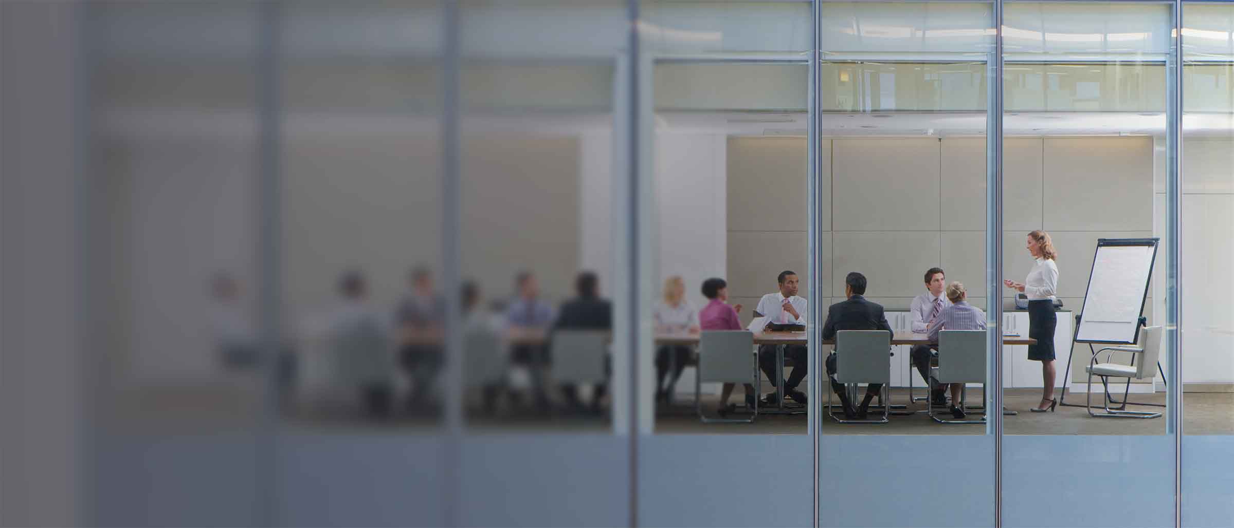 A person presents to colleagues seated at a conference table in a glass-windowed meeting room viewed from outside the building.