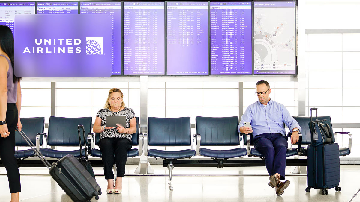 United Airlines logo in an airport terminal with passengers sitting in chairs near large digital flight information boards.